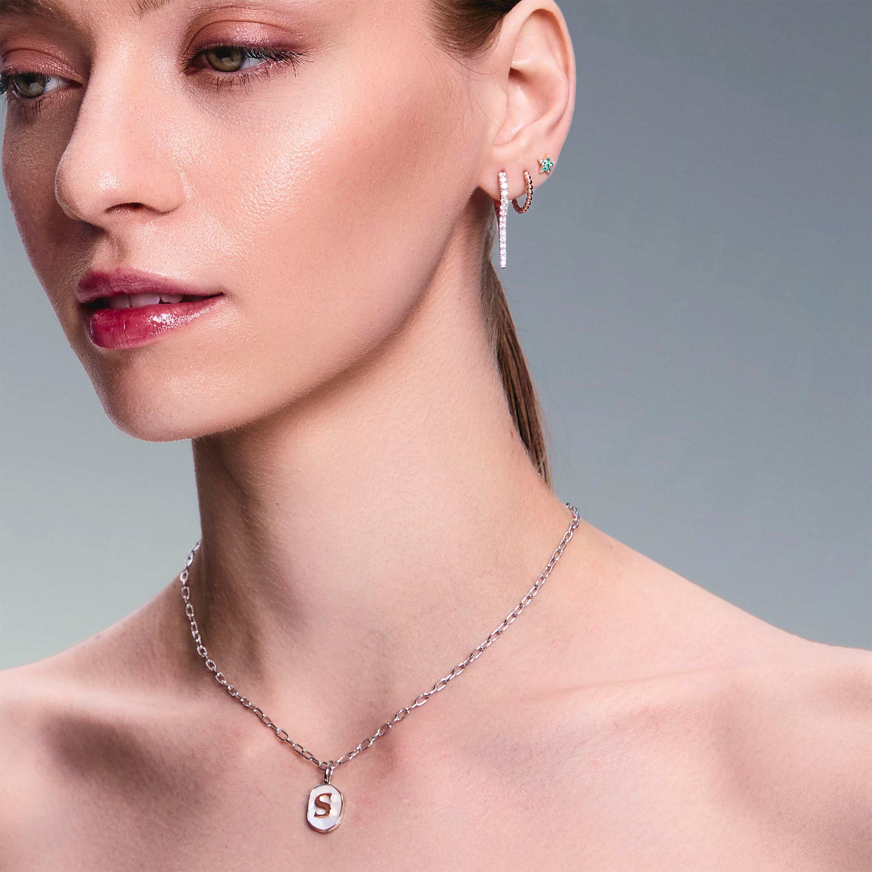 beautiful model wearing a mother of pearl pendant with a gold letter, diamonds drops earrings, gold hoops and star studs earrings. 