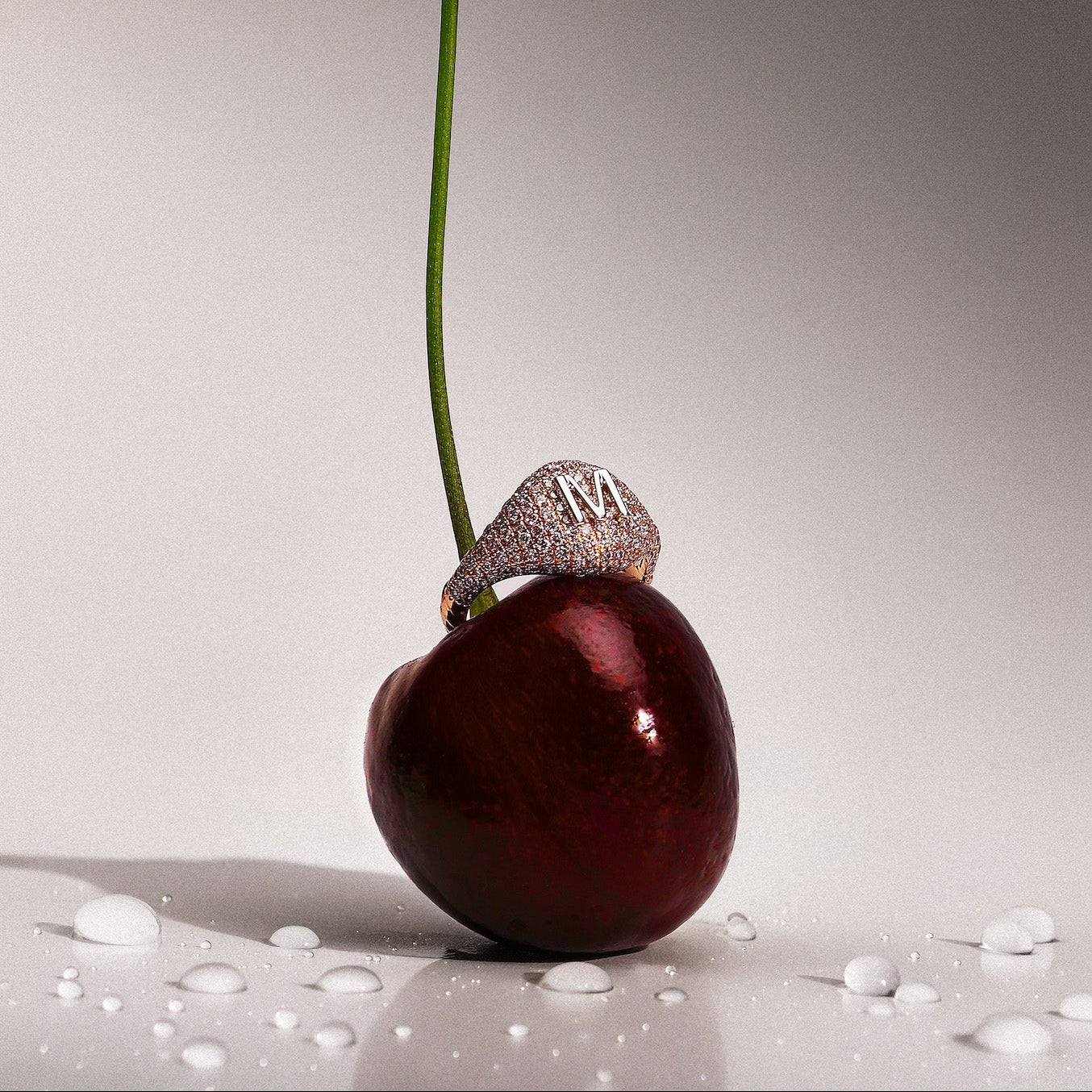 A dark red cherry with a long green stem stands upright on a glossy white surface with water droplets. A pave diamonds signet 14k gold ring with a large white gold letter 'M' sits on the cherry's stem.