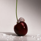 A dark red cherry with a long green stem stands upright on a glossy white surface with water droplets. A pave diamonds signet 14k gold ring with a large white gold letter 'M' sits on the cherry's stem.