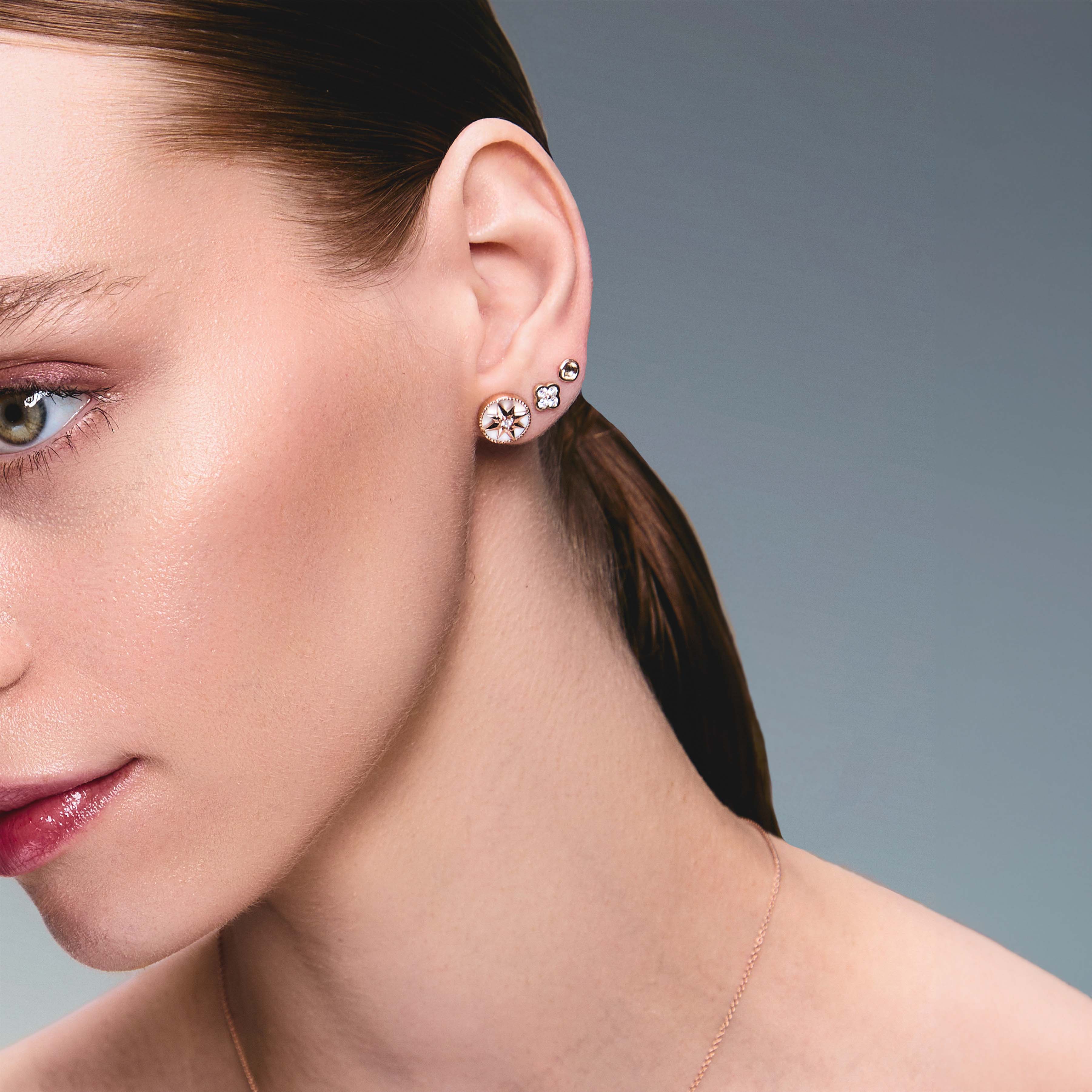 A pair of gold studs earrings with a simple design, resembling a prayer bowl or birds' nest, a clover diamonds earring, a mother of pearl lodestar earring, on an ear.