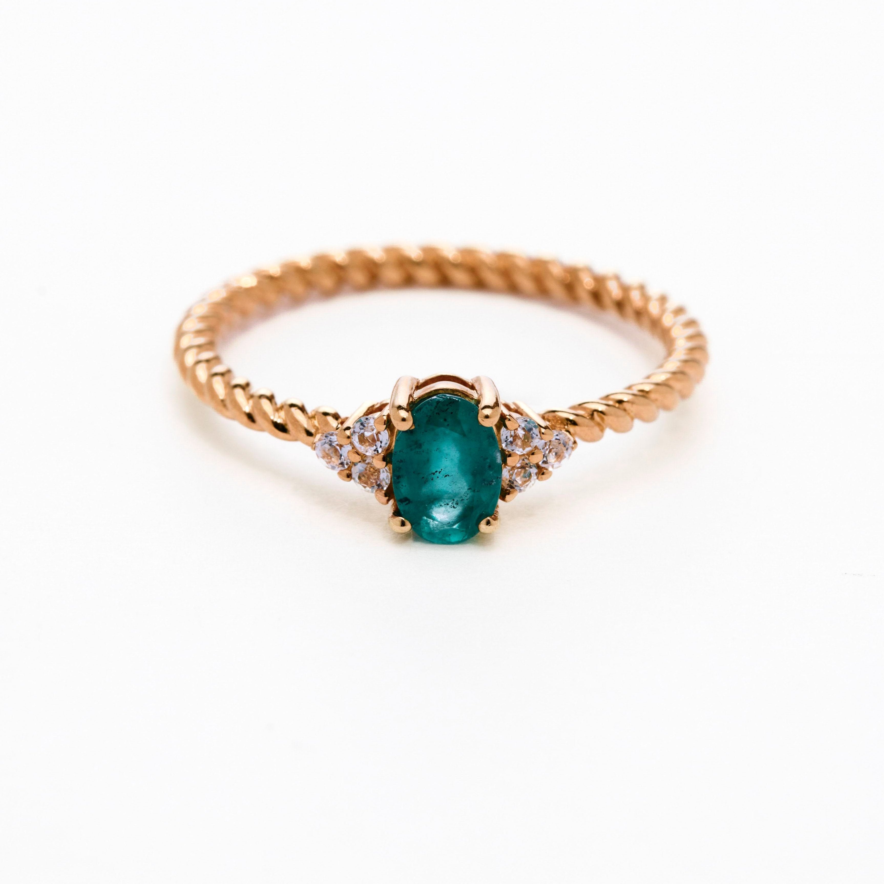 A rose gold ring featuring a central emerald stone flanked by white diamonds on either side, set against a white background.