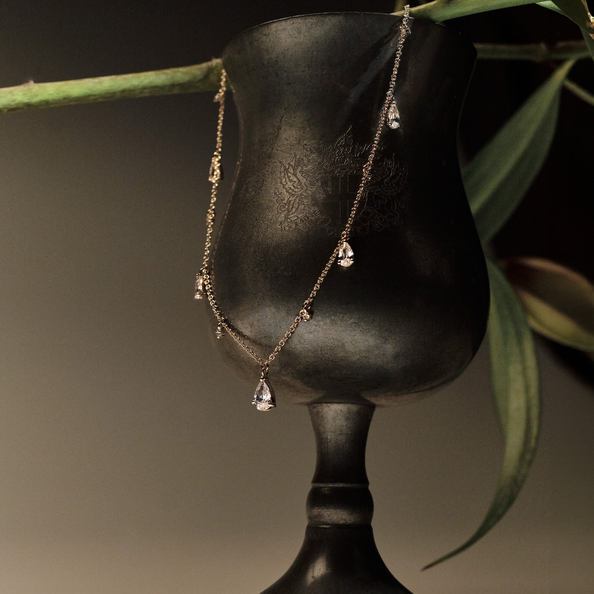 Gold Necklace with teardrop and round diamonds pendants on a black stand with green leaves in the background.