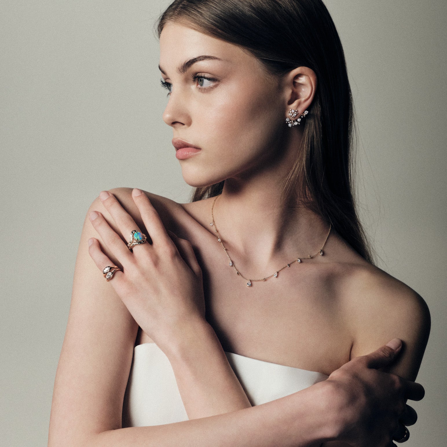 Woman wearing gold jewelry including bloom diamonds earrings, raindrops teardrops necklace, marquise diamonds signet pinky rings, and an australian opal and diamonds ring on a neutral background.