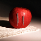 A red apple with two vertical rows of sparkling white gold earrings attached to its side, photographed on a white surface with a lace cloth and a dark background.