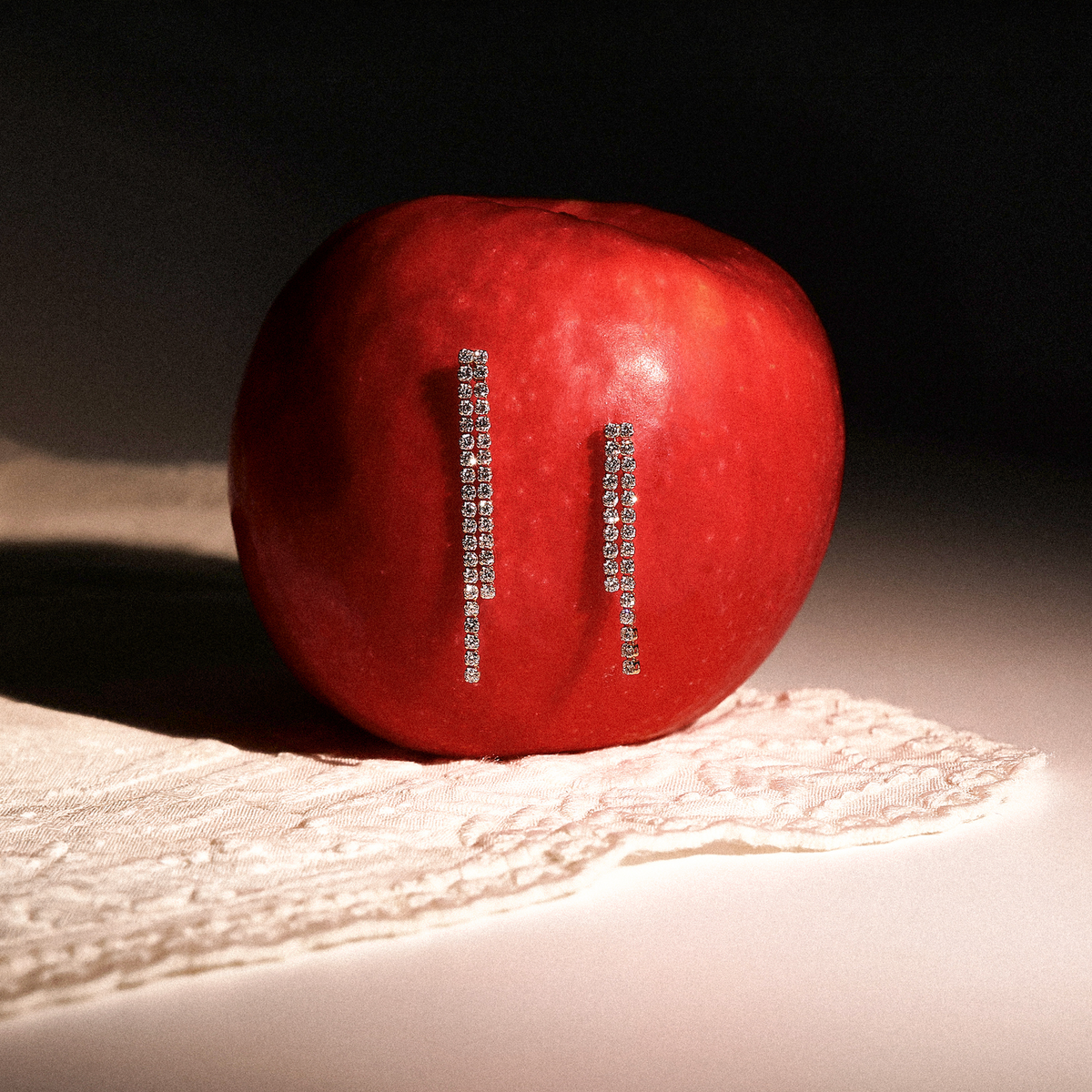 A red apple with two vertical rows of sparkling white gold earrings attached to its side, photographed on a white surface with a lace cloth and a dark background.