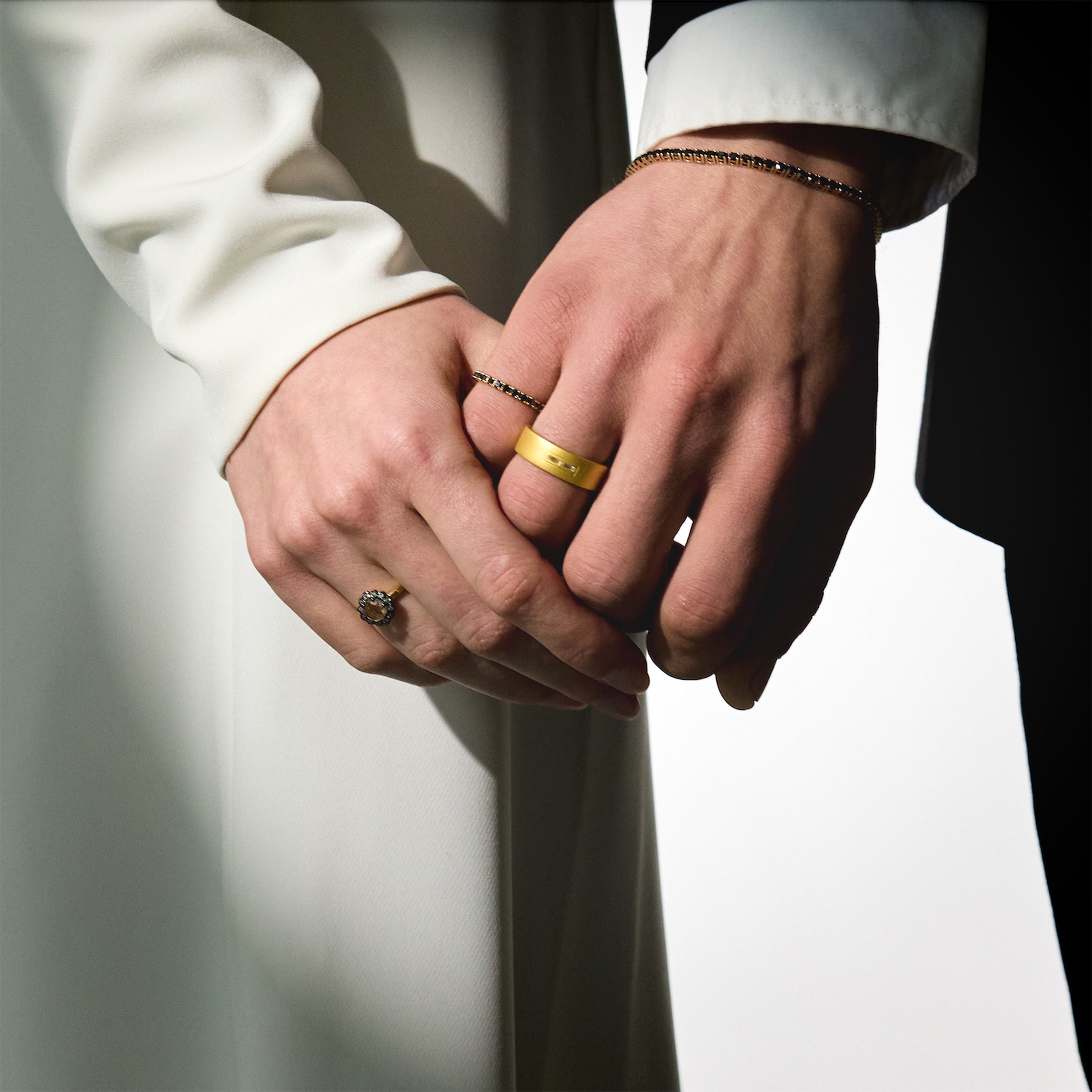 Two hands holding each other, wearing gold wedding rings, black diamonds tennis bracelet and ring, on a neutral background