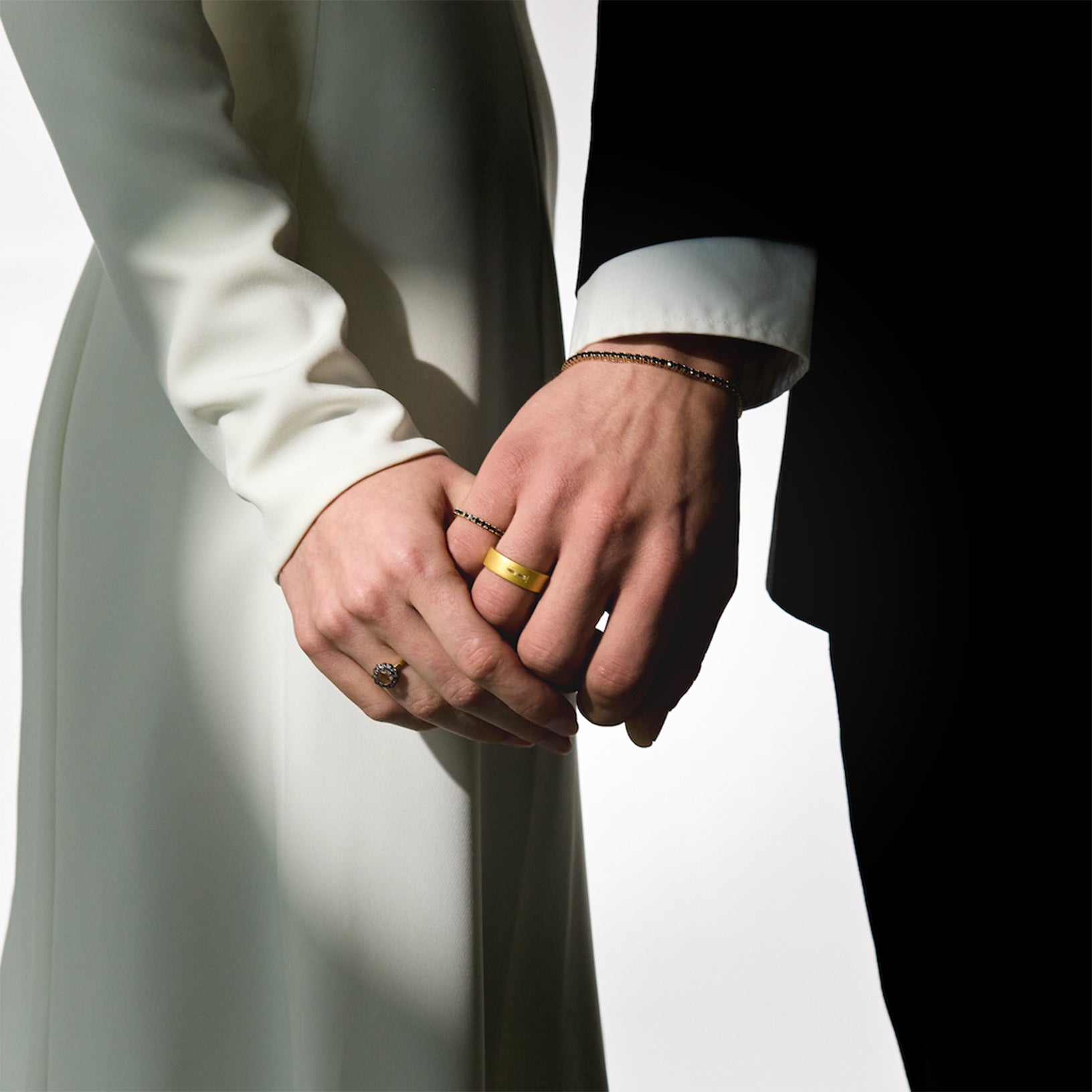 Two hands holding each other, wearing gold wedding rings, black diamonds tennis bracelet and ring, on a neutral background