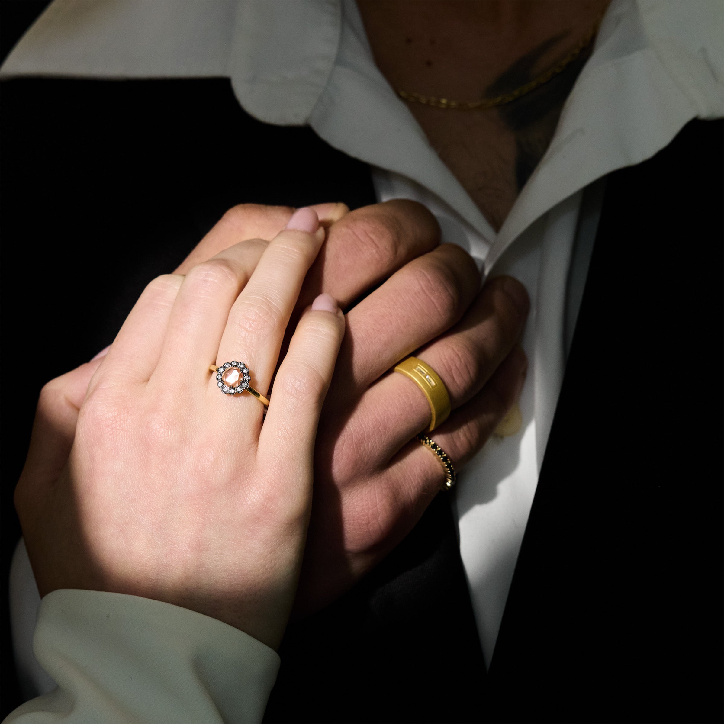 Two hands holding each other, wearing gold wedding rings, black diamonds tennis bracelet and ring, on a neutral background