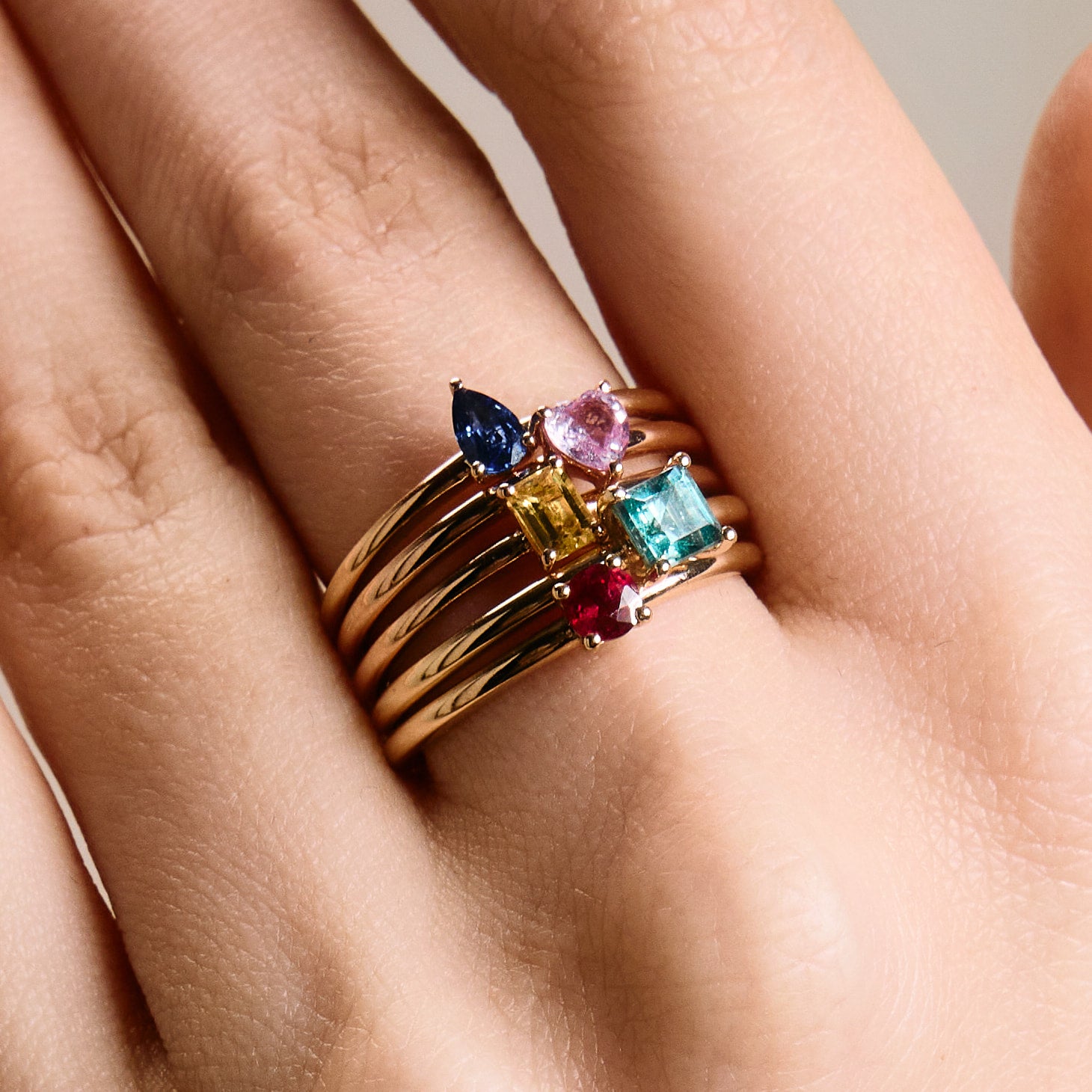 A collection of gold rings with a central ruby stone, accompanied by smaller multicolored stones, stacked on a person's finger.