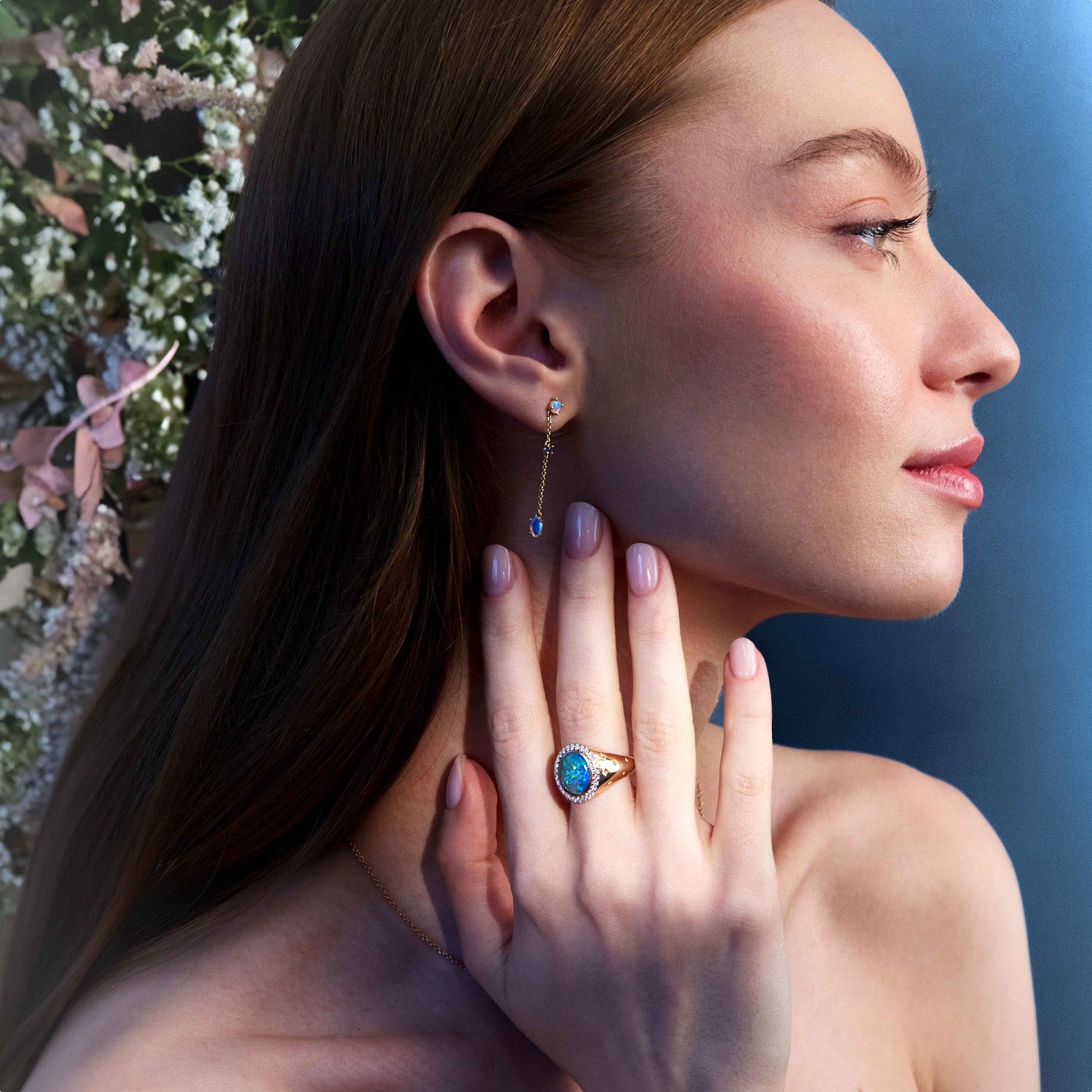 A person wearing a pair of gold earrings with round blue or green sapphires and Australian Opals, and an Australian Oval Opal Signet ring, standing near a floral backdrop.