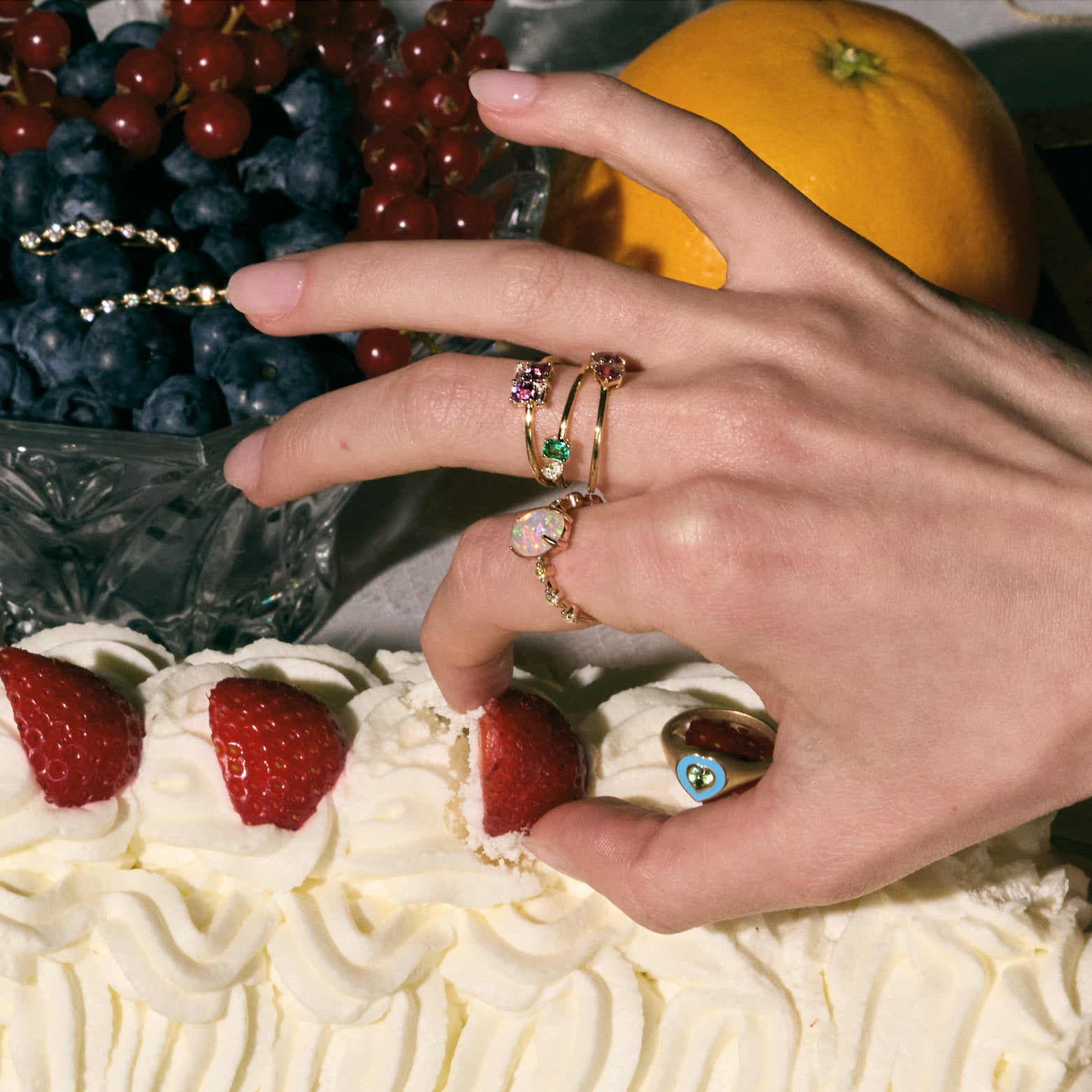 Hand with rings on a cake decorated with strawberries and cream, surrounded by fruits.