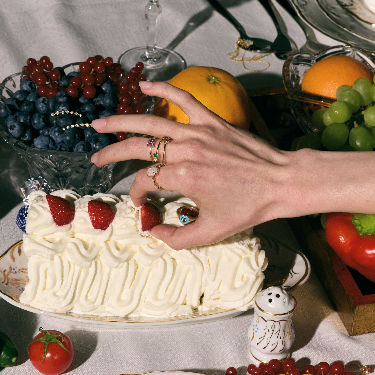 Hand wearing multiple gemstones rings - emerald, sapphire, diamonds - reaching towards a cake with fruit on a table. 