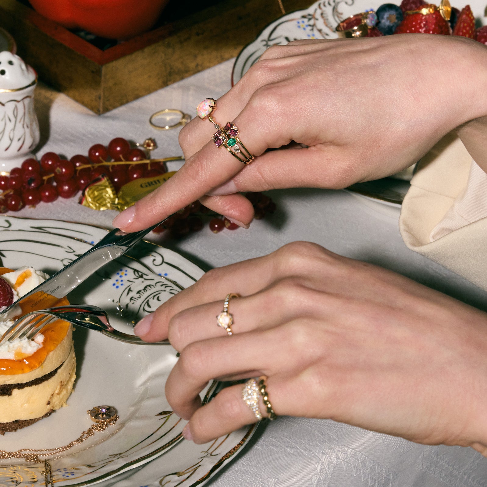 Close-up of hands with rings cutting a dessert on a plate with a decorative edge, Wearing multiple gold rings, adorned with diamonds, emeralds and sapphire. 