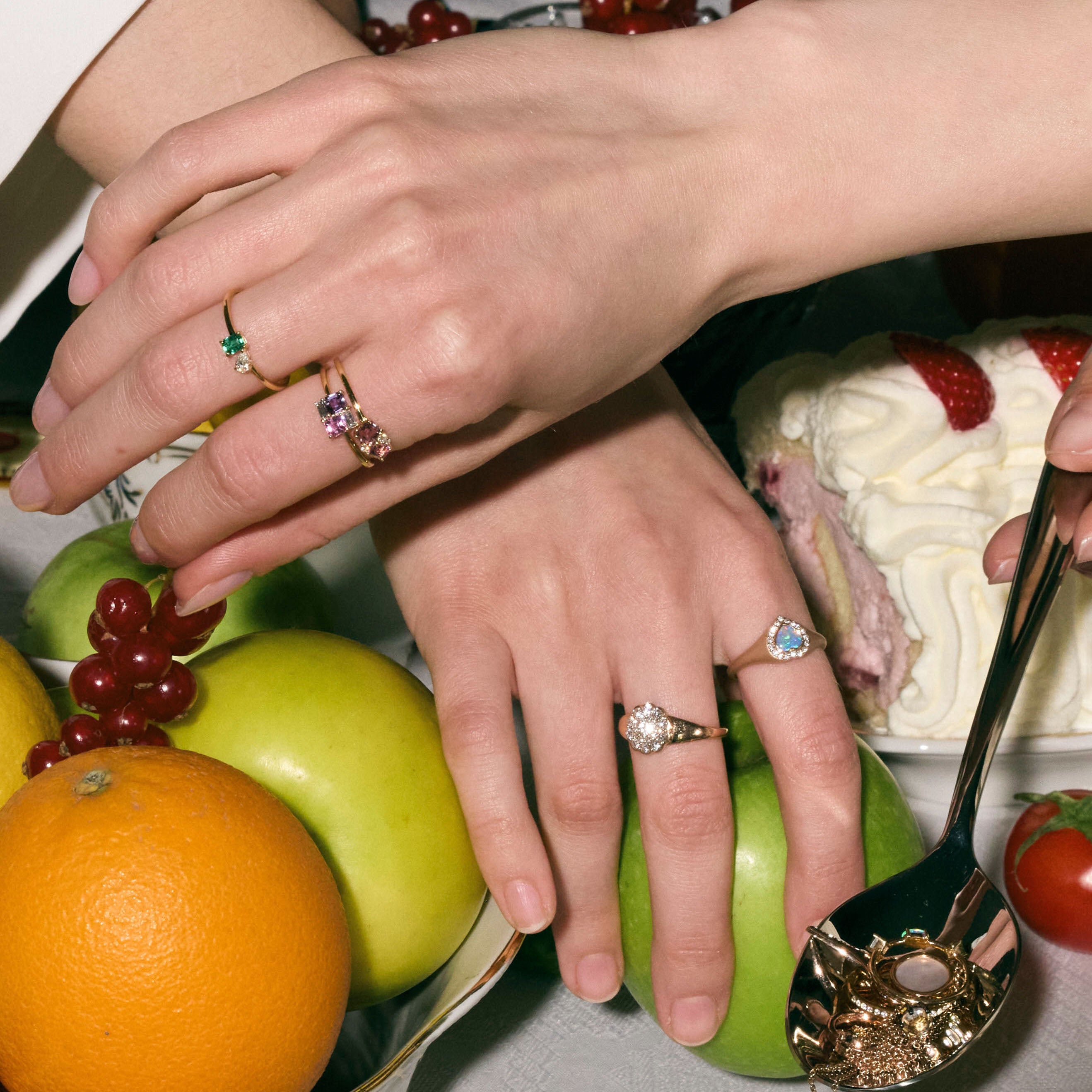 Close-up of hands with rings and different gemstones - diamonds, australian opal heart, emeralds, sapphires - on a table with fruits and a cake.