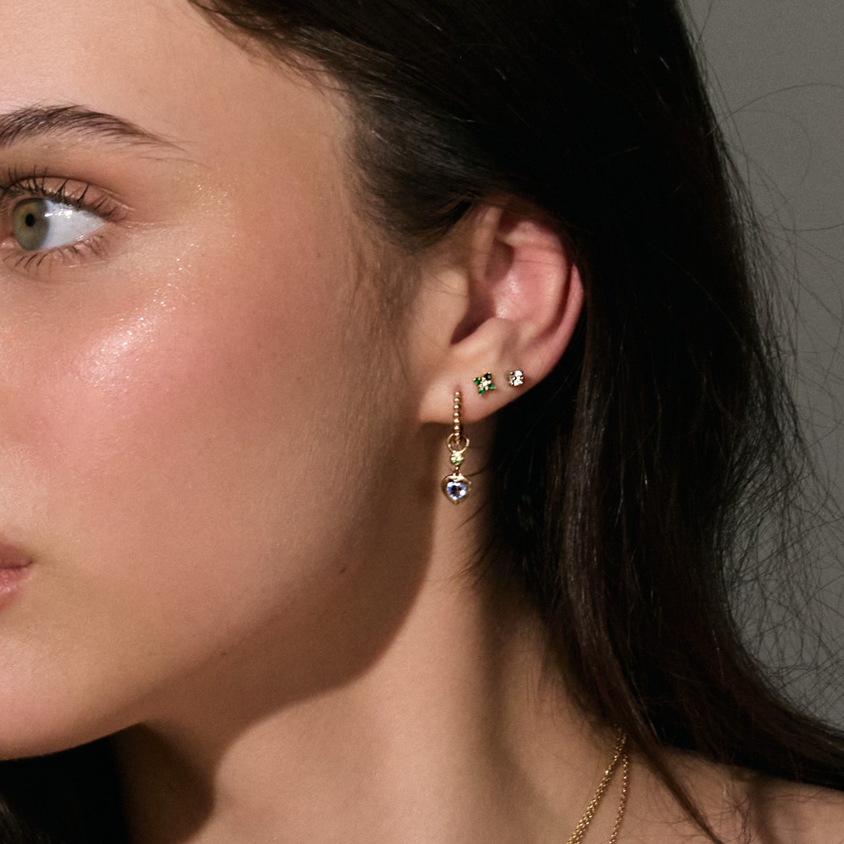 Close-up of a woman wearing stud 0,20cts diamond earrings, emerald earrings and hoop earrings with a neutral background