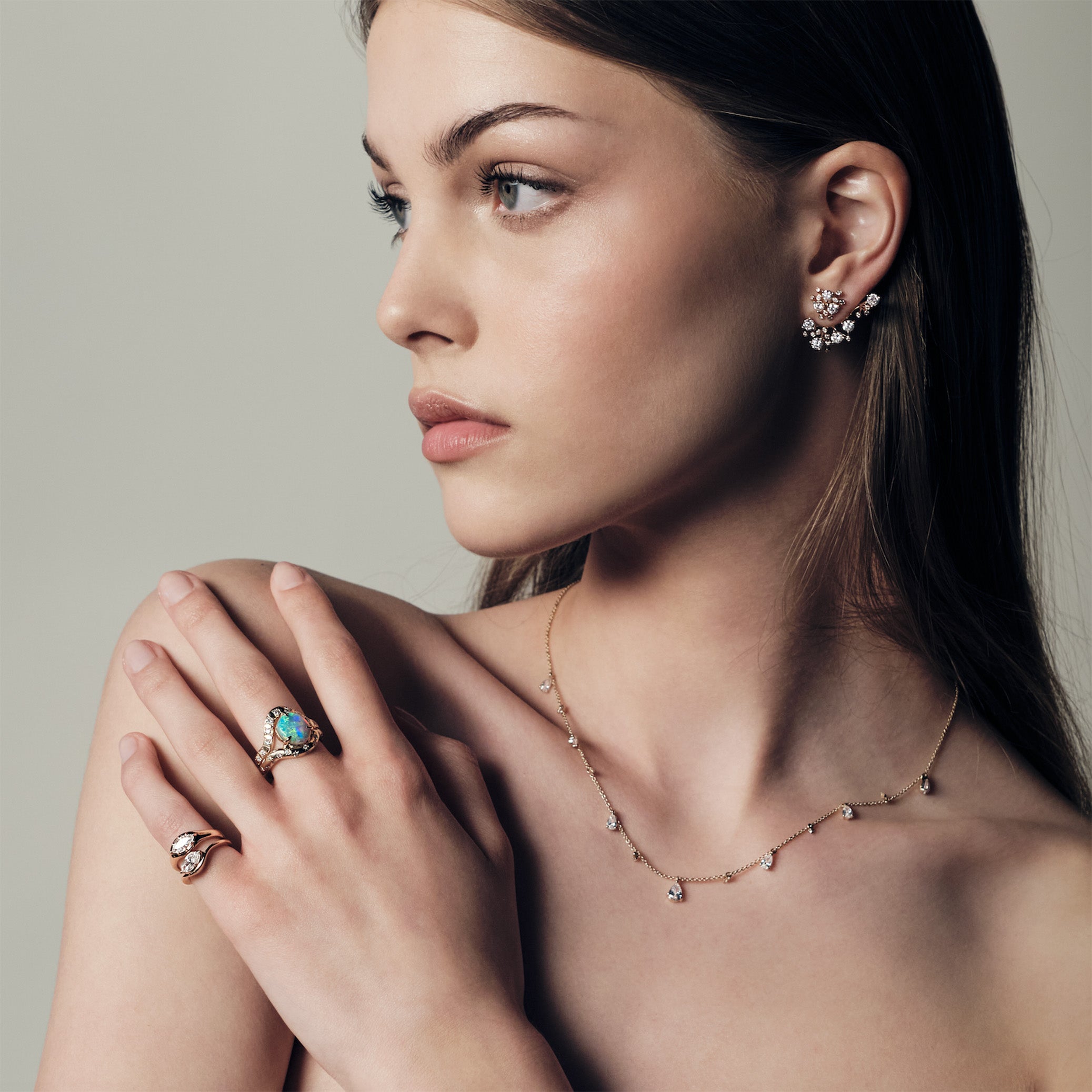 A woman wearing a ring with a australian opal and diamonds, two pinky ring with marquise diamonds set in curvy rose gold bands. She uis also wearing a teardrop diaomonds necklace and a cluster diamonds earcrawler earring. 