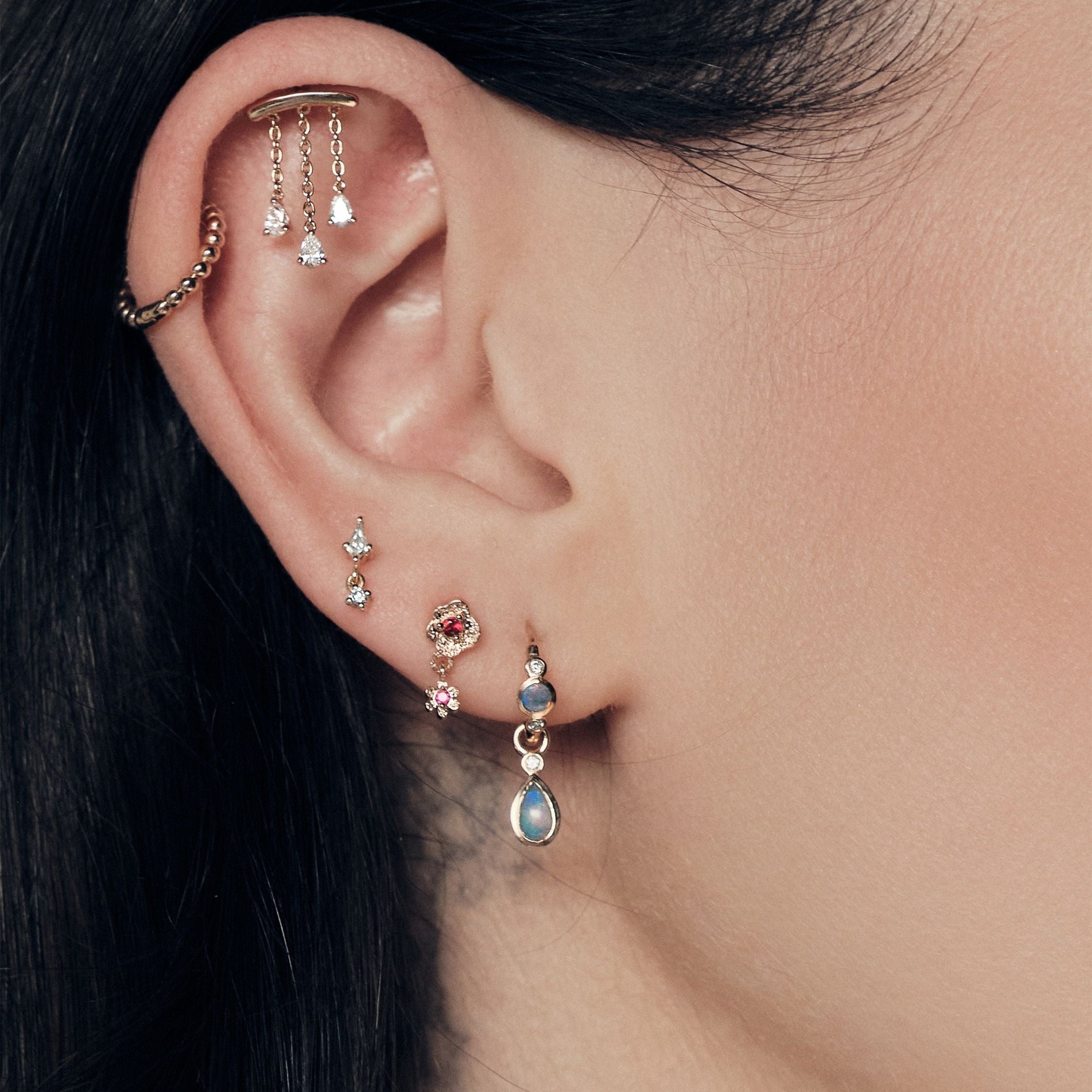 Close-up of an ear with multiple earrings on a blurred background. Chandelier diamonds stud, bubble hoop, Floating Kite Diamond stud, Wild Rose Stud With Ruby, Tiny Dew Drops Hoops and Teardrop Australian Opals Earrings accessories. 