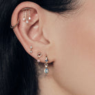 Close-up of an ear with multiple earrings on a blurred background. Chandelier diamonds stud, bubble hoop, Floating Kite Diamond stud, Wild Rose Stud With Ruby, Tiny Dew Drops Hoops and Teardrop Australian Opals Earrings accessories. 