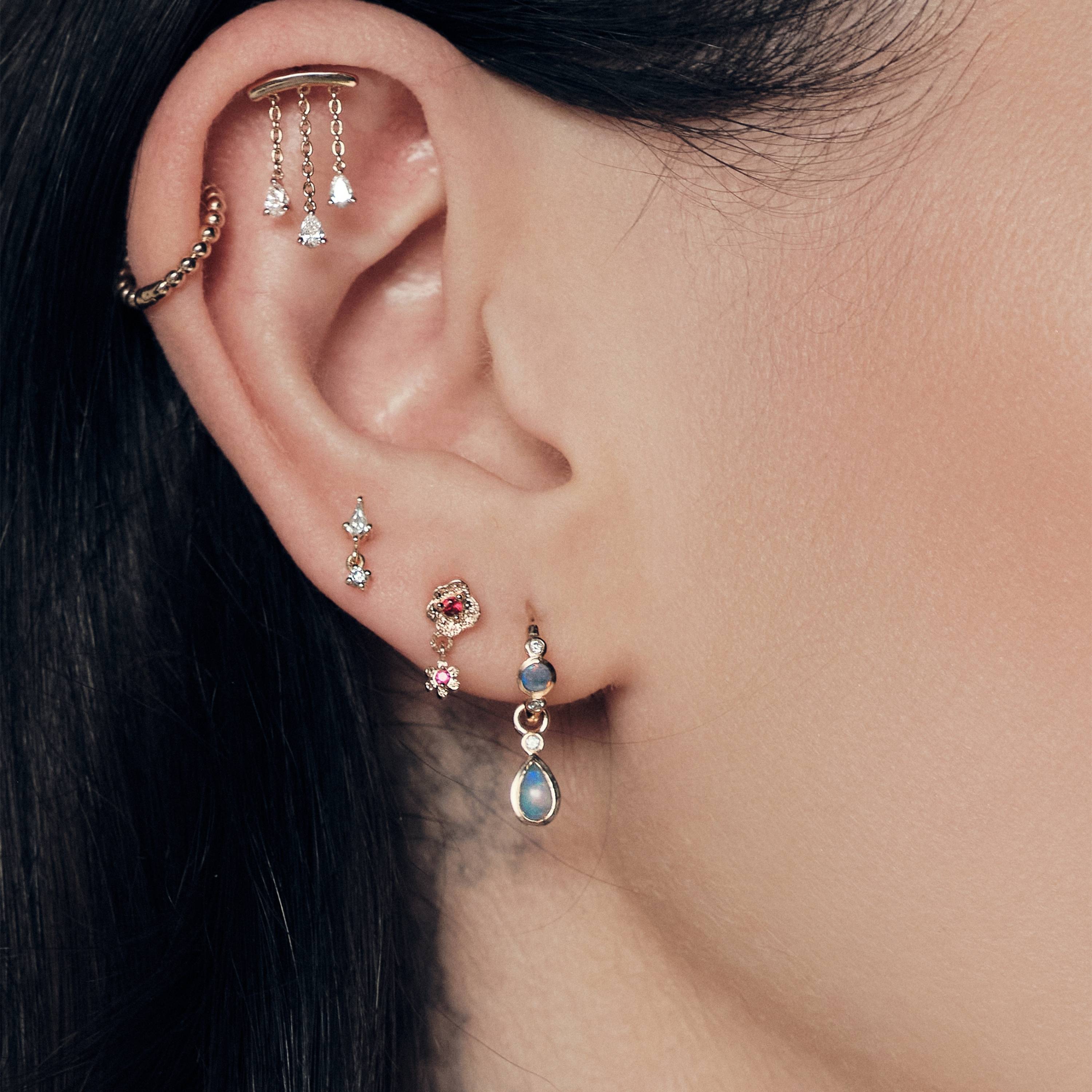 Close-up of an ear with The Chandelier Stud, Bubble Time 13mm Hoops, Floating Kite Diamond Stud, The Wild Rose Ruby Stud, Tiny Dew Drops Australian Opal Hoops, focusing on jewelry details.