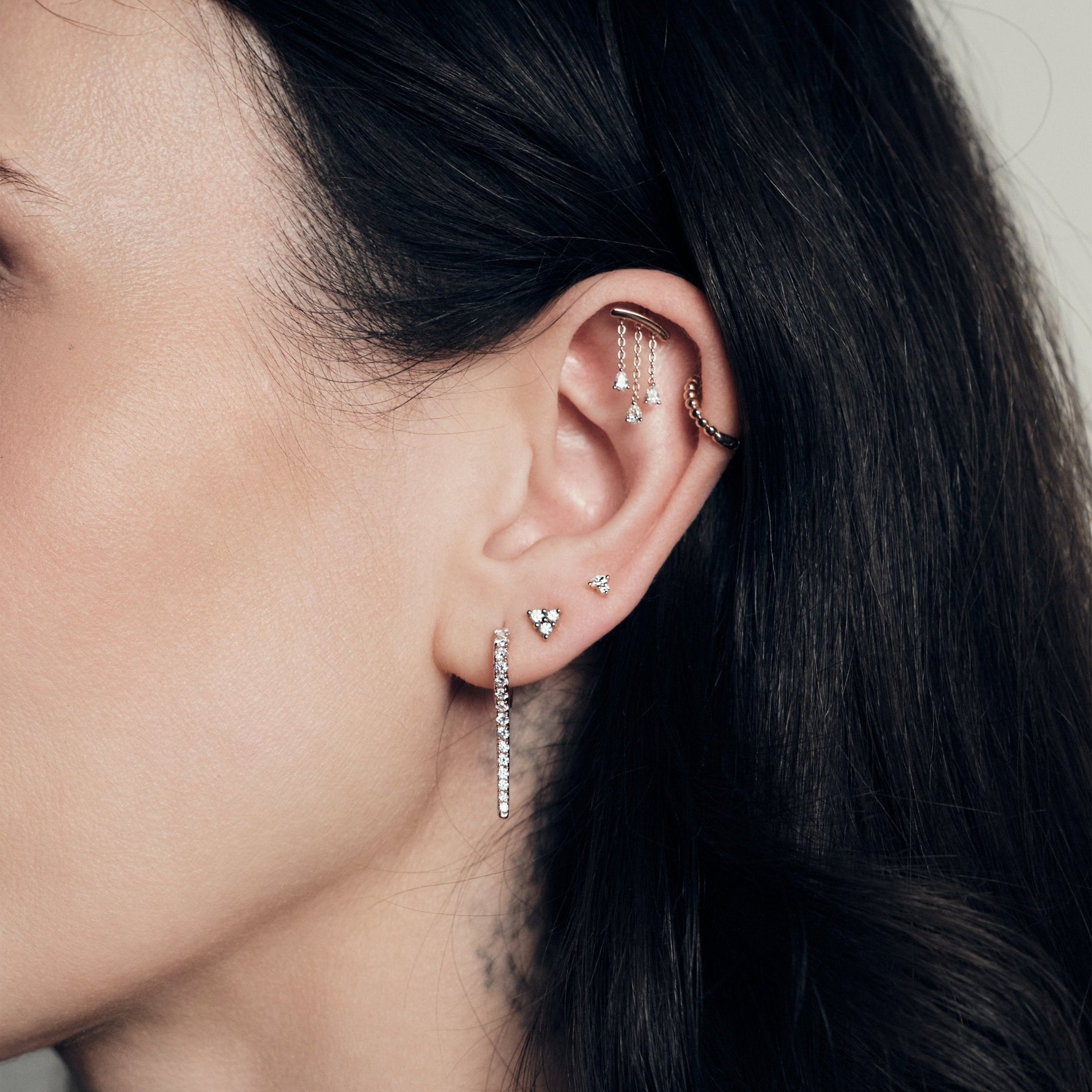 Close-up of an ear with multiple earrings, chandelier teardrop diamonds stud, bubble time 13mm hoop, marquise and round diamond stud, dot dot dot, icicle earring, against a neutral background