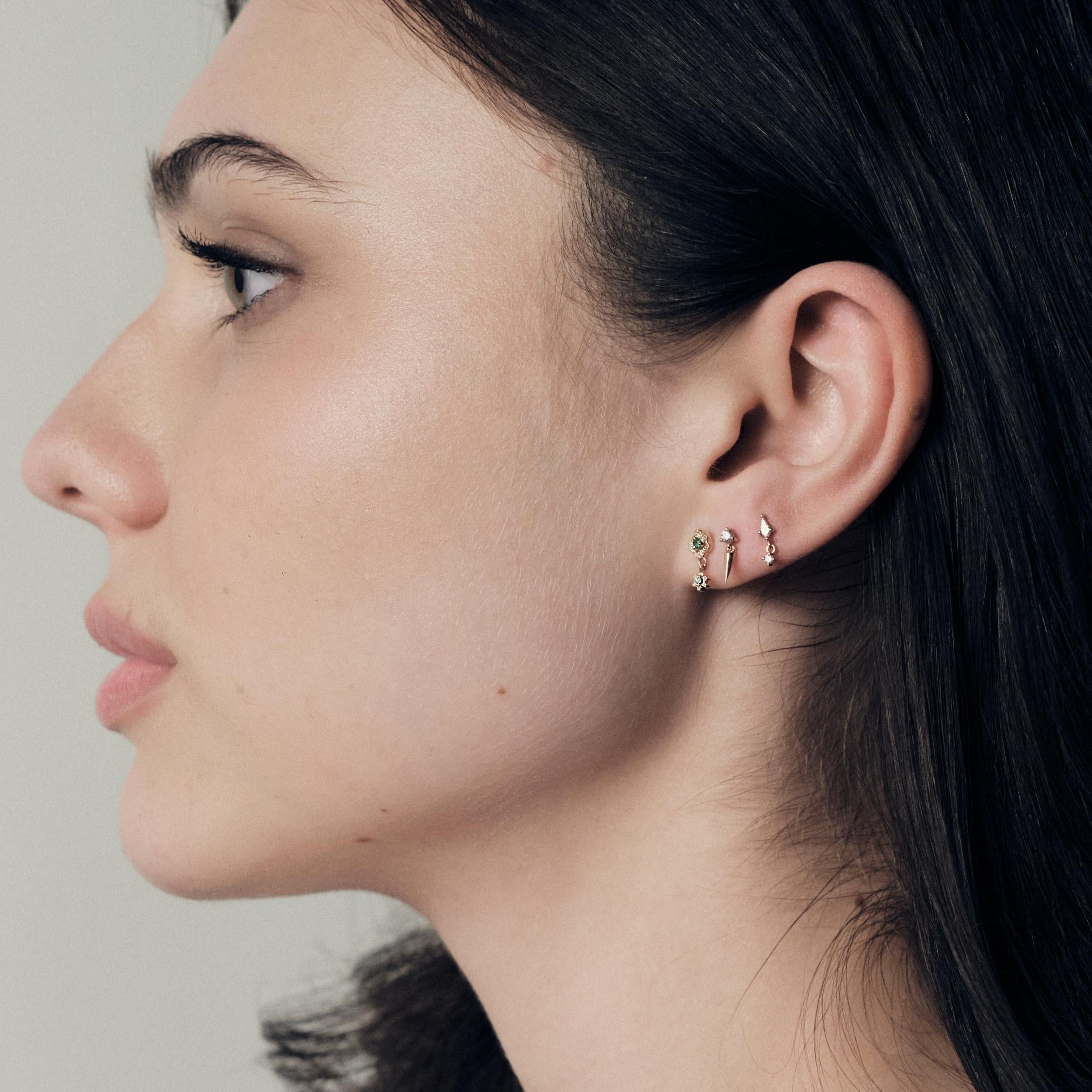 Close-up of a woman's profile wearing earrings on a neutral background. She is wearing Wild Rose Gold studs with emeralds, Floating Gold icicle Stud and Floating Kite Stud madeof yellow gold. 