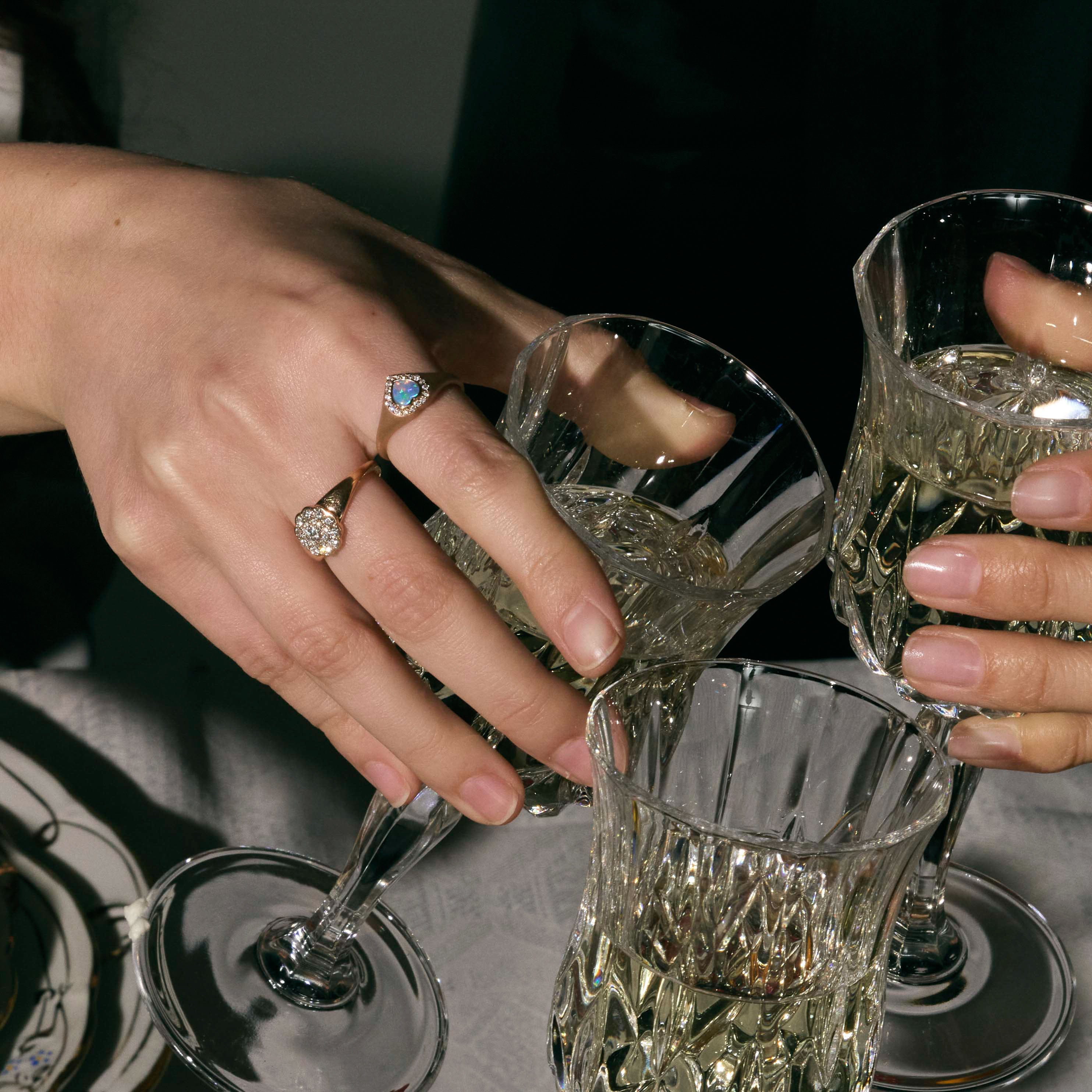 Two hands with gold, australian heart oals and diamonds rings holding crystal glasses filled with a clear liquid on a dark surface.
