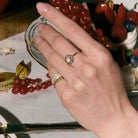Hand wearing rings with diamonds, and pinky rings on a table with fruit and jewelry.