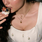 Woman eating a dessert full of gold jewelry, wearing gold necklaces with various pendants, cherry flower pendant, bee pendant and heart pendant.