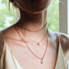 Close-up of a person wearing three rose gold necklaces withe diamonds and a diamond flower pendant, with a blurred natural background